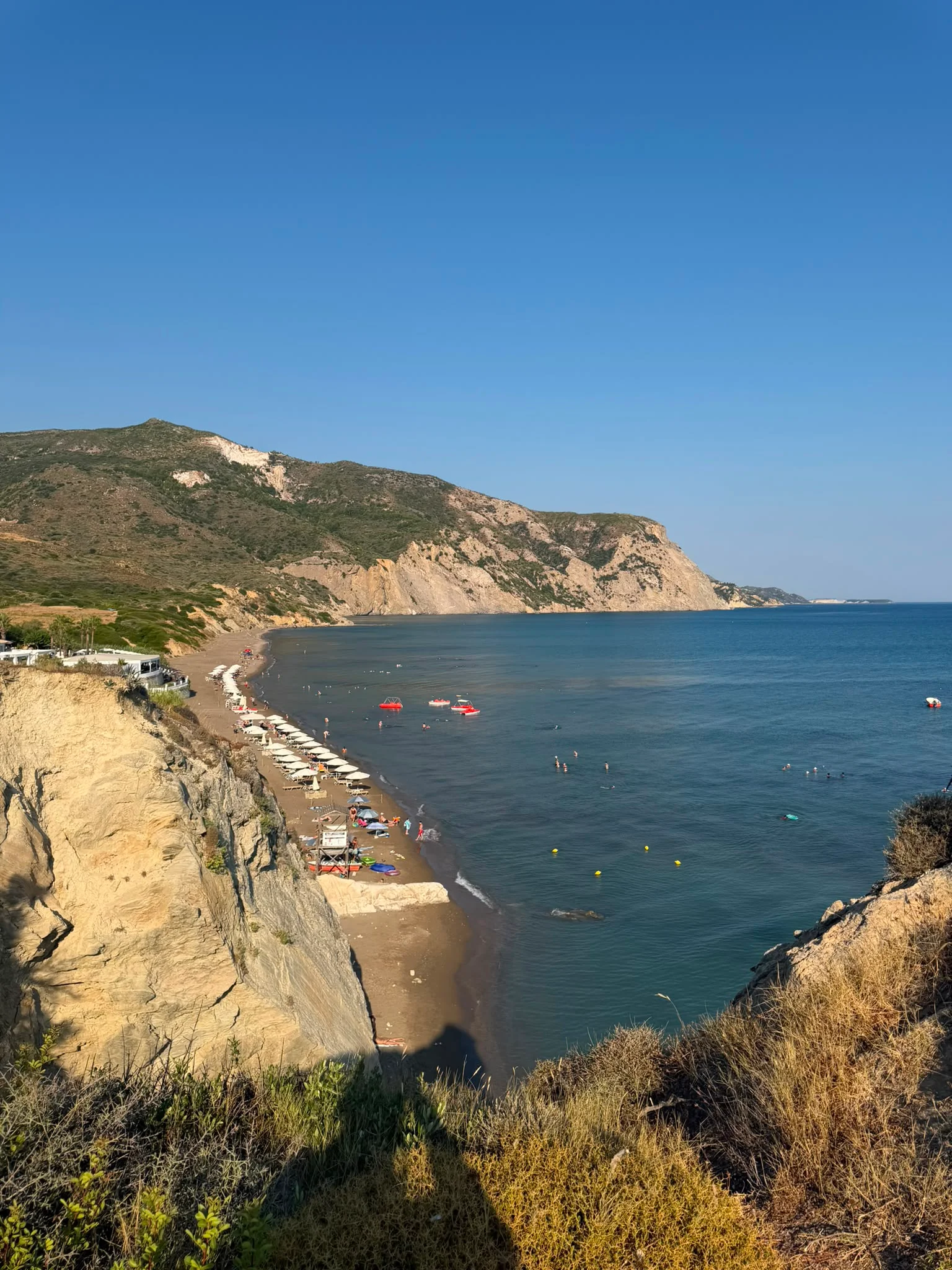 Crystal Beach seen from the cliff path, with rows of sunbeds along the shore and boats moored offshore